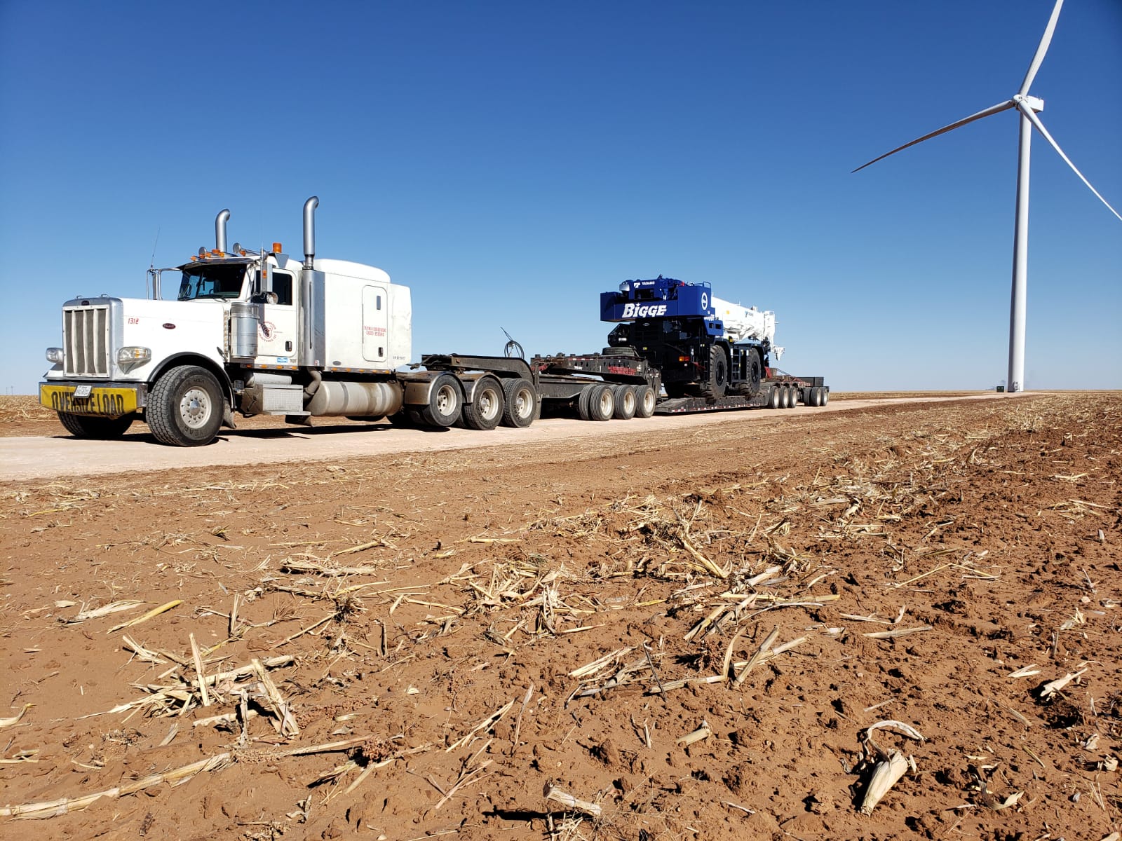 White Peterbilt with lowboy trailer hauling a Bigge rough terrain crane at a wind farm